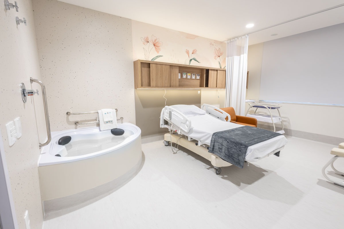 Fotografia de quarto hospitalar, com banheira, leito, berço, poltrona e decoração floral suave.