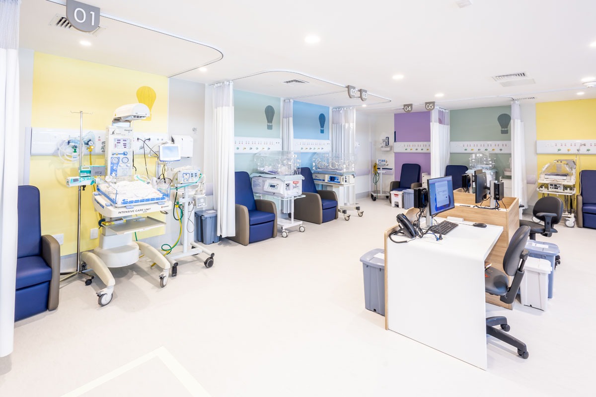 Fotografia de UTI neonatal, com incubadoras, equipamentos médicos, poltronas azuis e posto de trabalho.