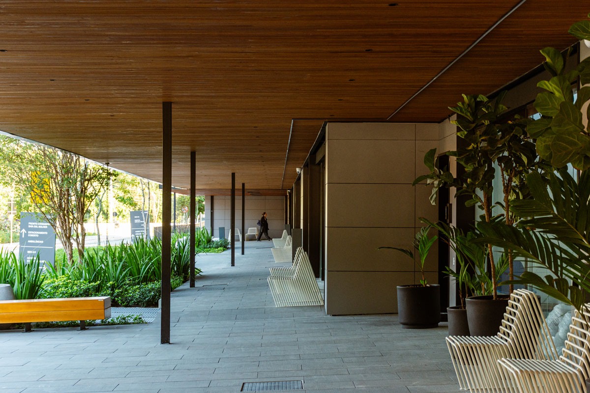 Fotografia de corredor coberto, com plantas, bancos, cadeiras brancas e teto revestido de madeira.