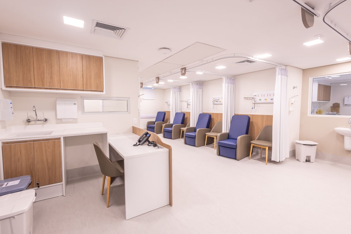 Fotografia de sala de atendimento hospitalar, com poltronas azuis, divisórias com cortinas e bancada de apoio.
