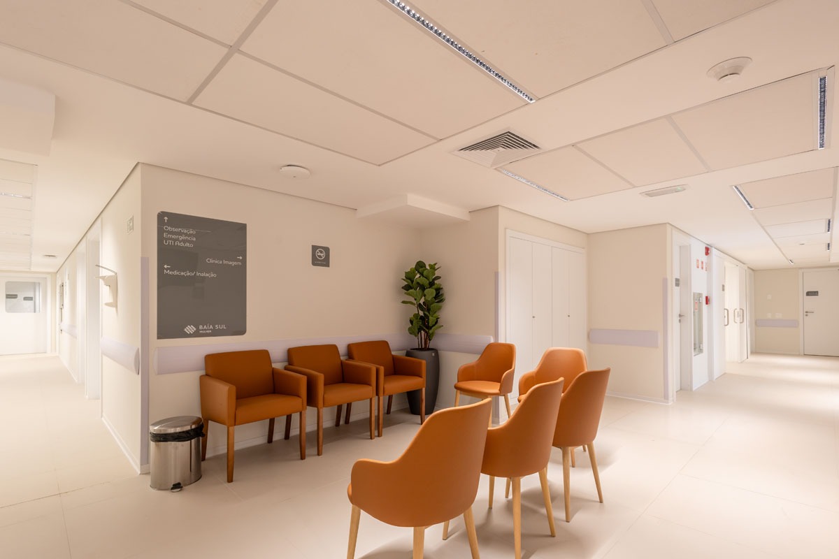 Fotografia de sala de espera hospitalar, com cadeiras laranja, placa de orientação e corredor iluminado.v
