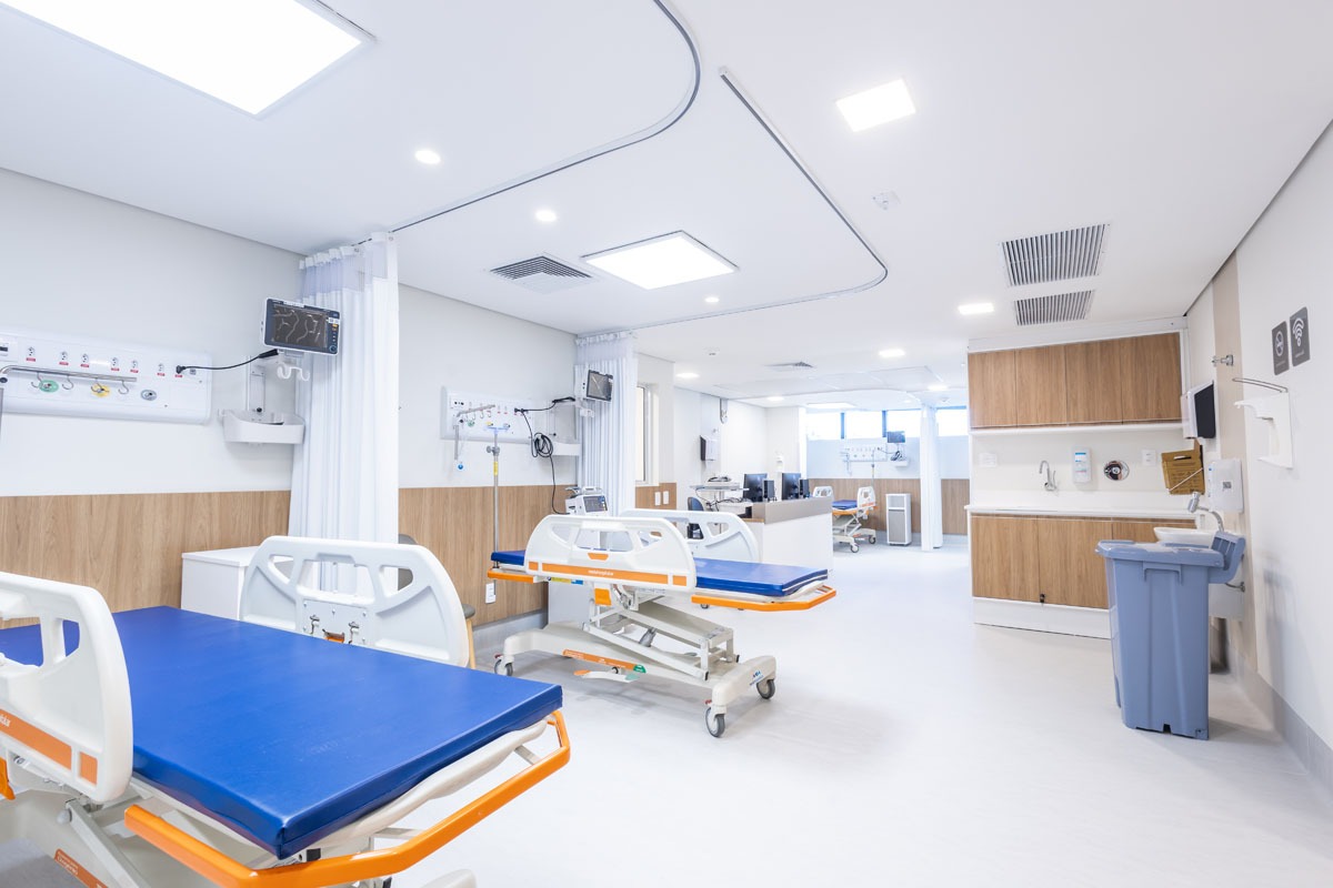 Fotografia de unidade hospitalar com leitos, equipamentos médicos, cortinas de privacidade e armários.