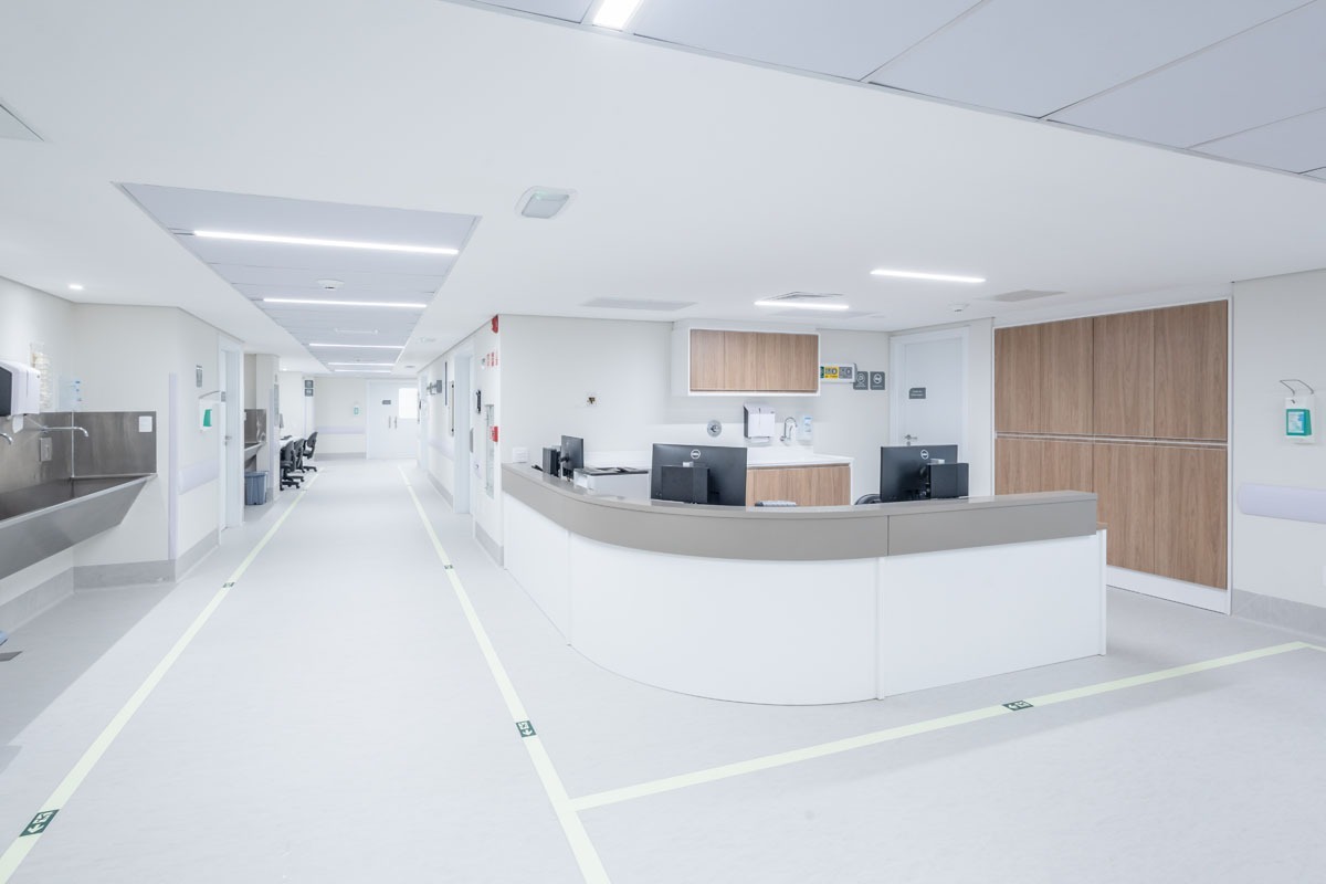 Fotografia de posto de enfermagem em corredor hospitalar, com balcão, computadores e área branca iluminada.