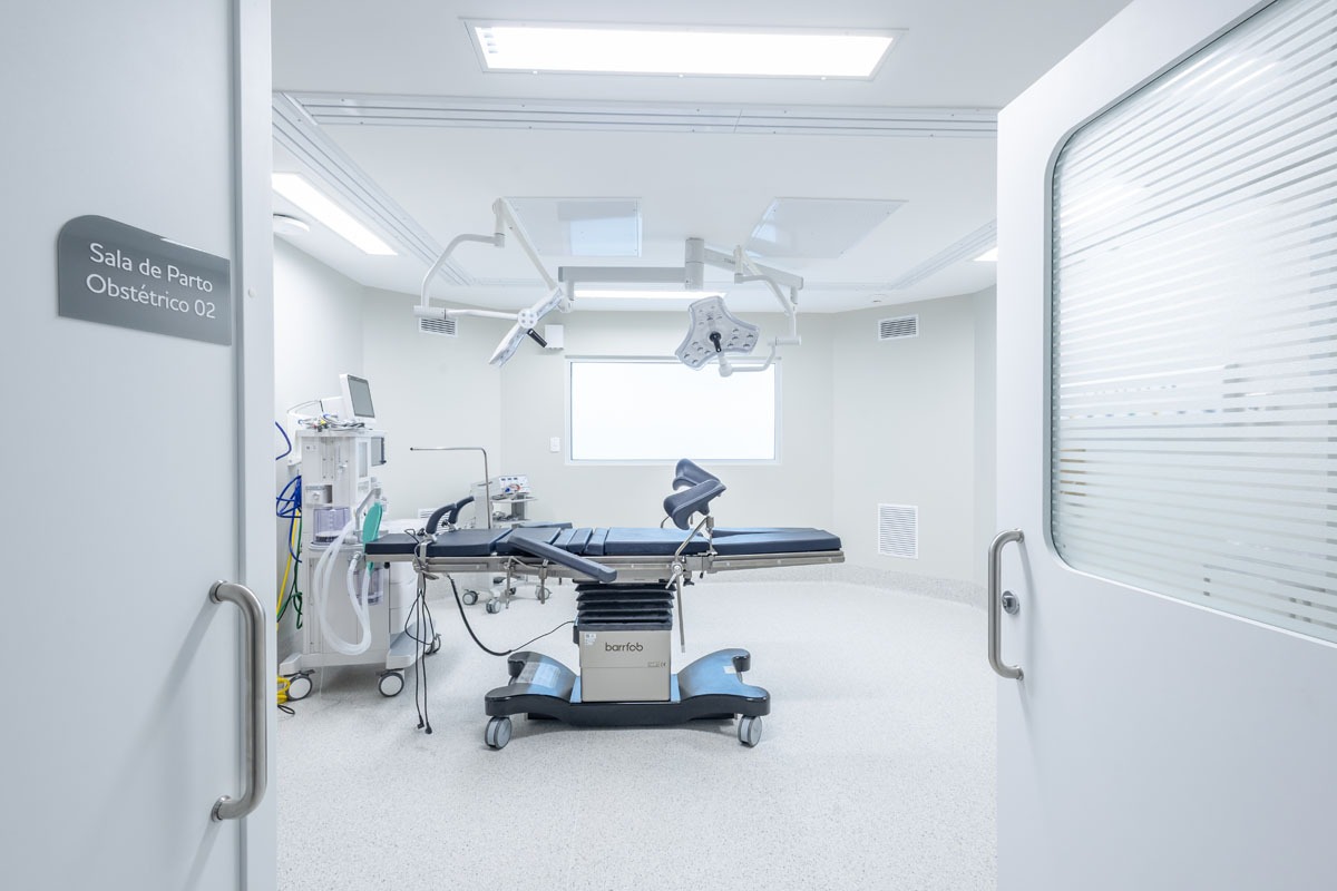 Fotografia de sala de parto obstétrico, com maca, equipamentos médicos e iluminação cirúrgica.