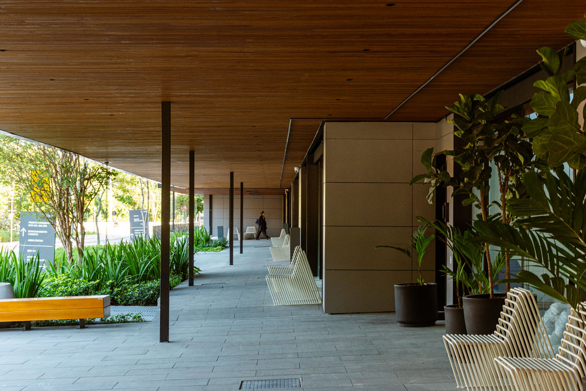 Fotografia de corredor coberto, com plantas, bancos, cadeiras brancas e teto revestido de madeira.