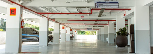 Fotografia de garagem coberta, com vagas, carros estacionados, sinalização e tubulação vermelha no teto.