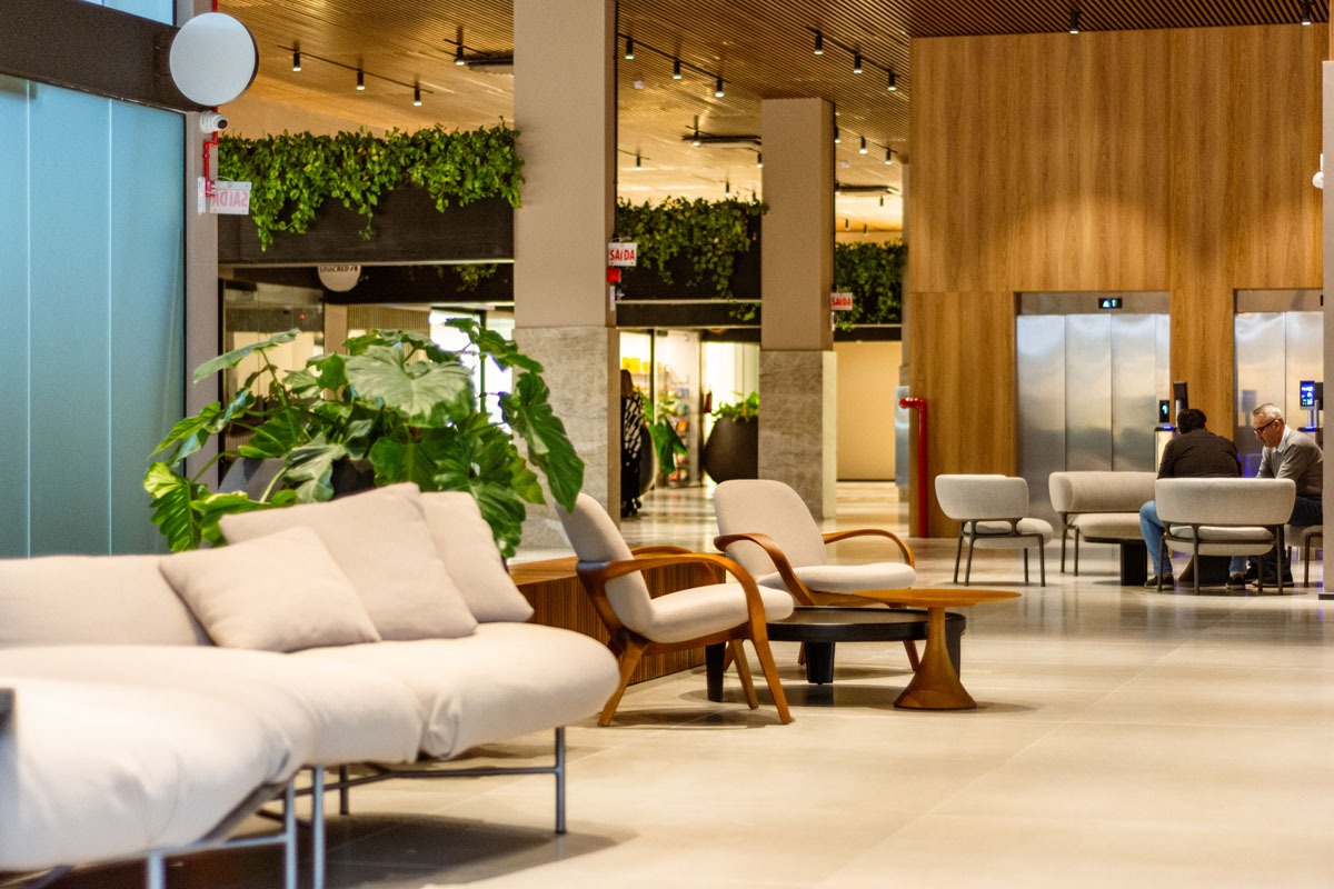 Fotografia de lounge moderno, com sofá, poltronas, plantas, elevadores e duas pessoas conversando ao fundo.